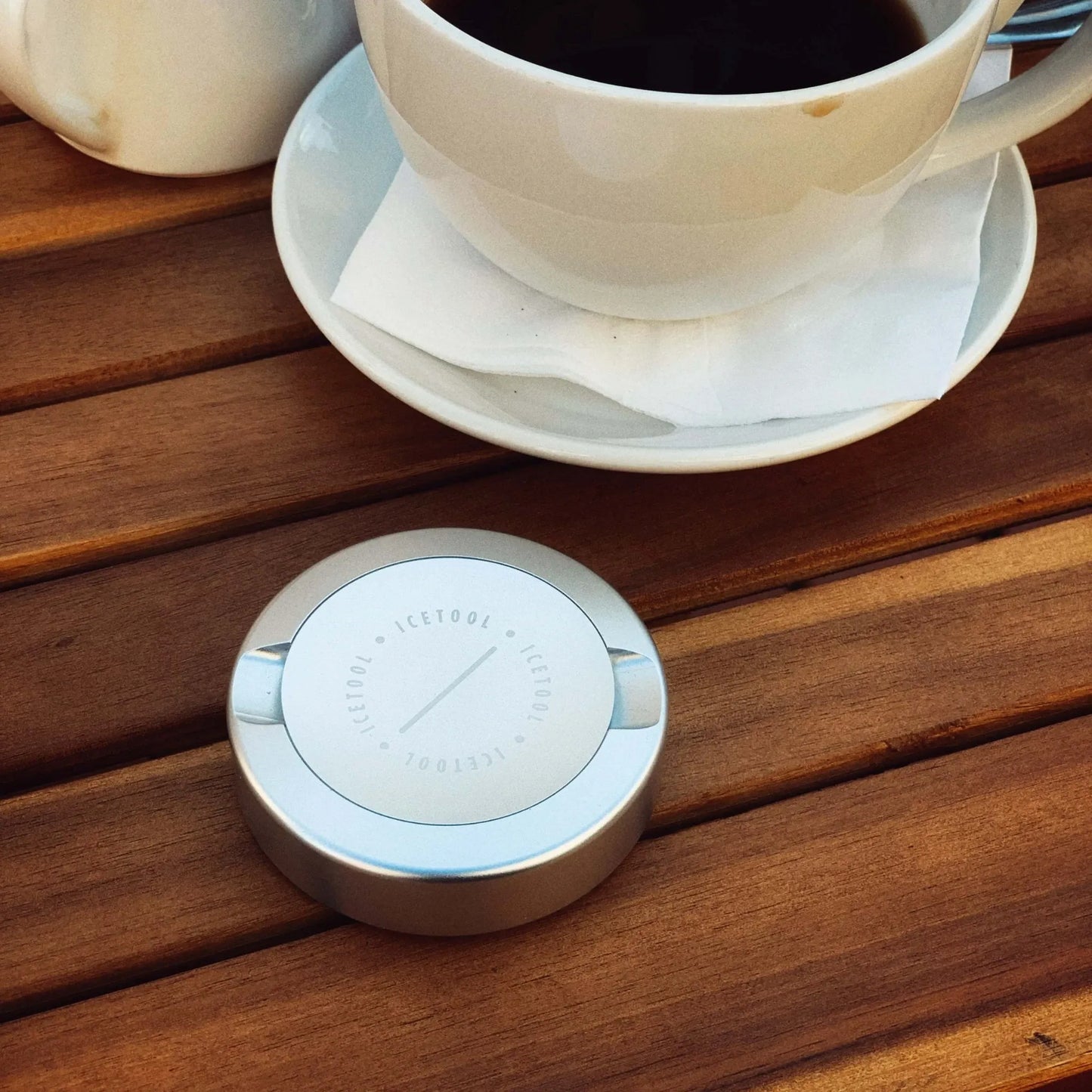 Metallic silver circular container with Icetool branding next to a cup of black coffee on a wooden table.