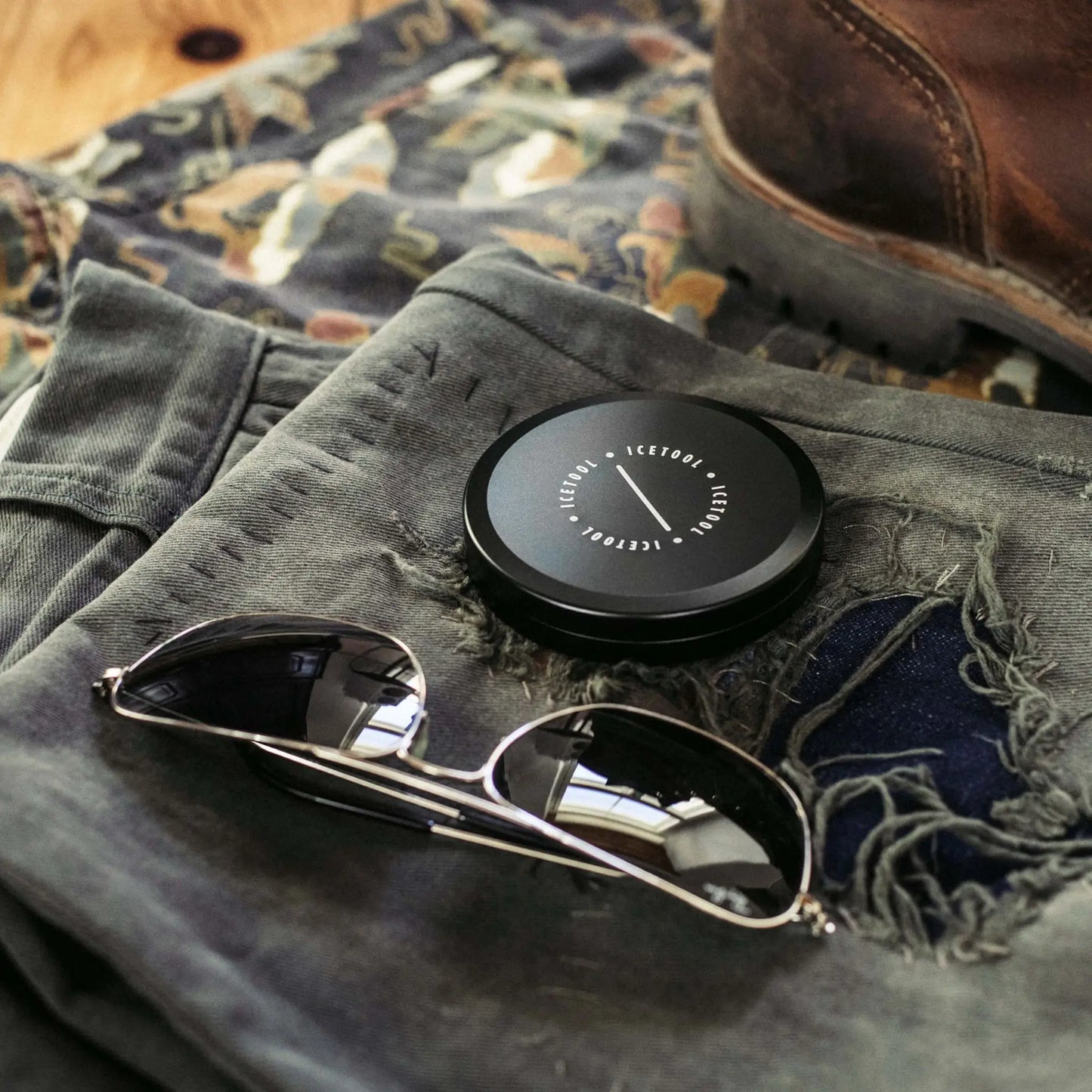 Black circular container with Icetool branding on a background with sunglasses, dark jeans and leather boots.
