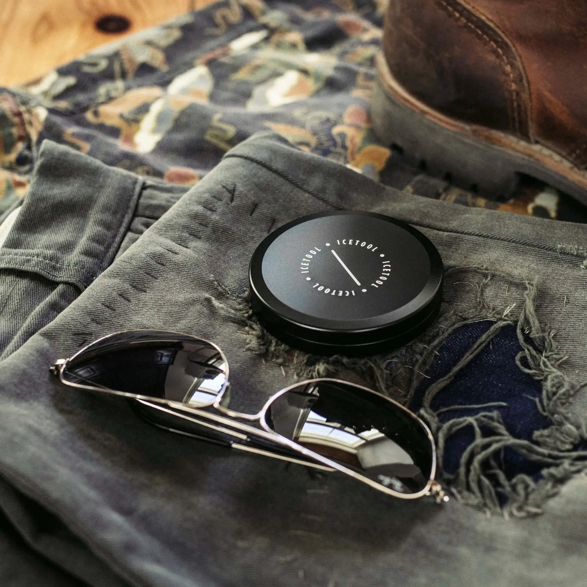 Black circular container with Icetool branding on a background with sunglasses, dark jeans and leather boots.