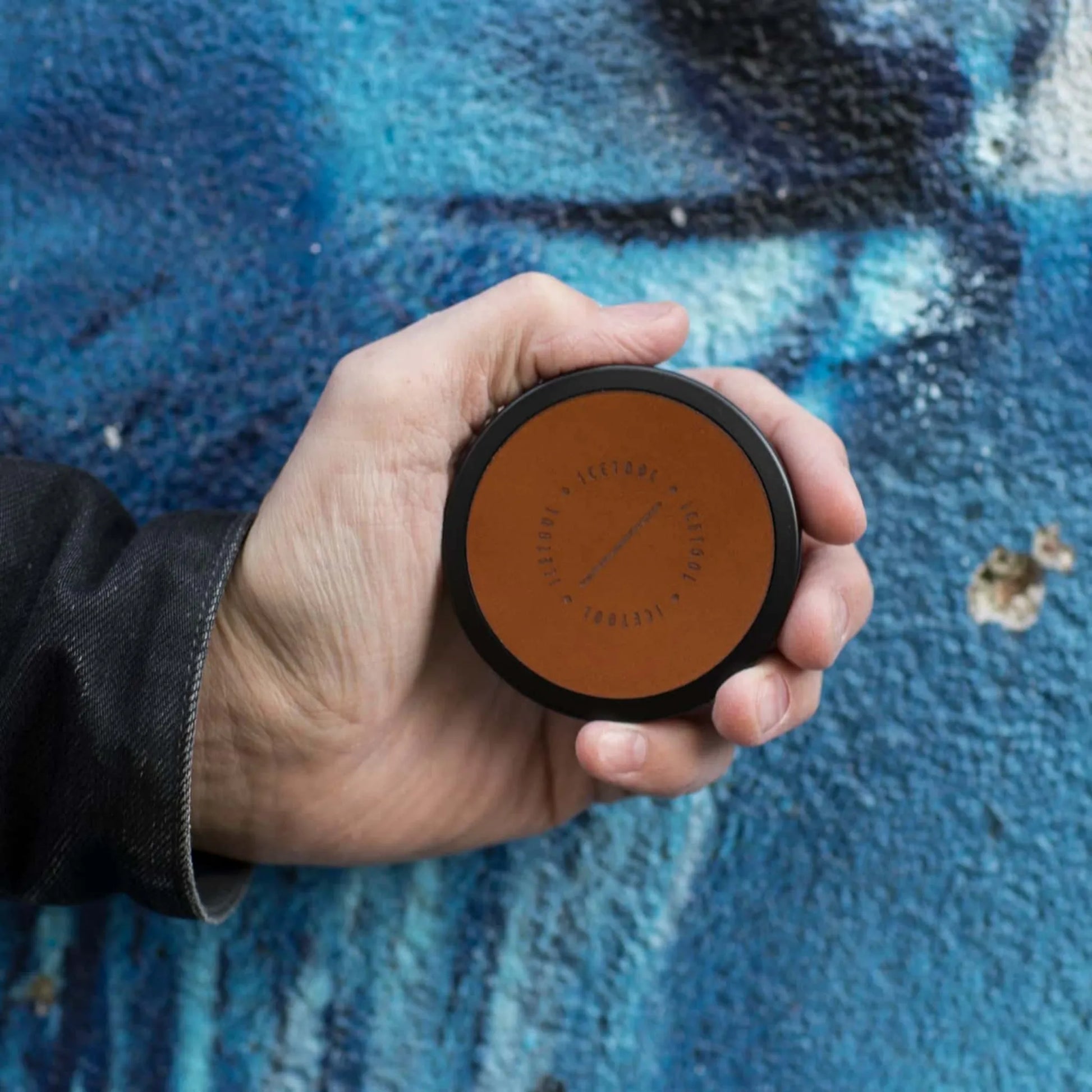 Metal and leather circular container with Icetool branding, being held in a hand against a blue wall.