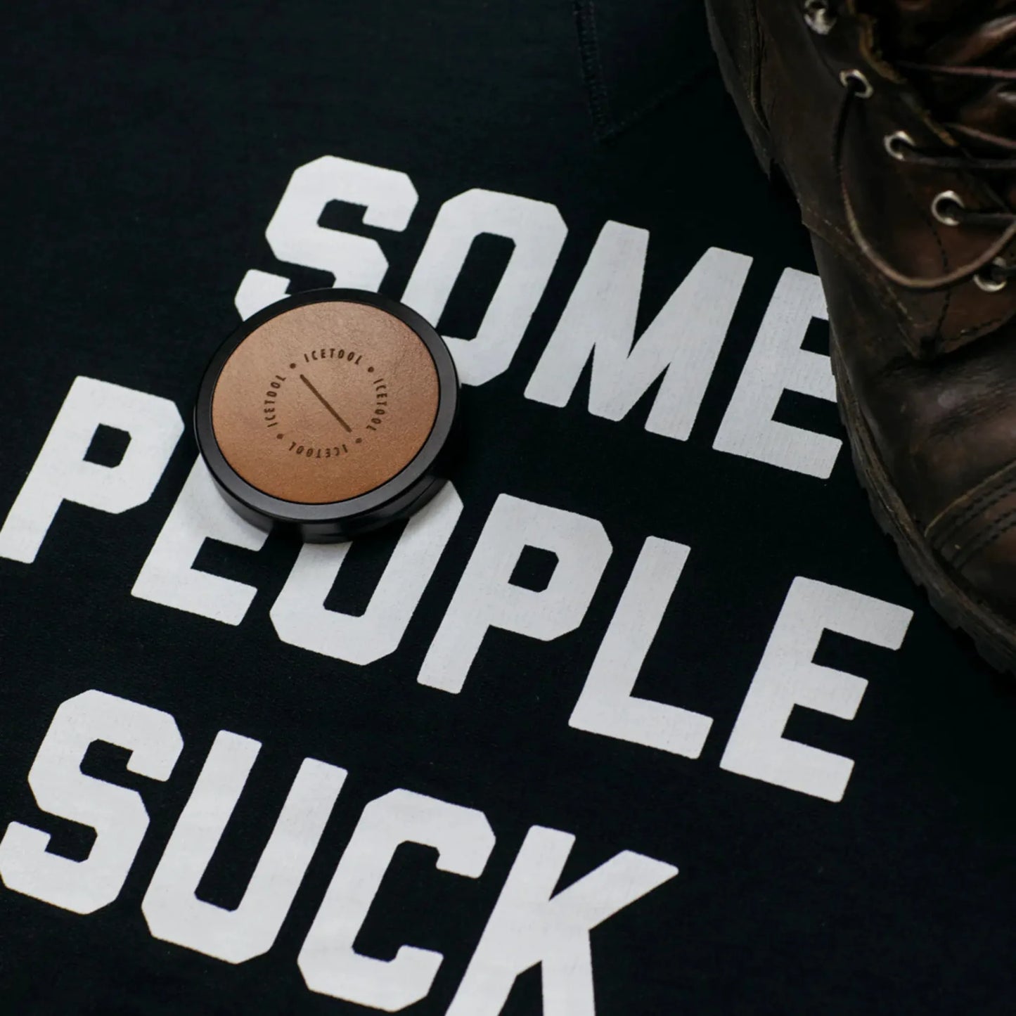 Black t-shirt with 'Some people suck' text, brown circular container, and brown boot on a black background