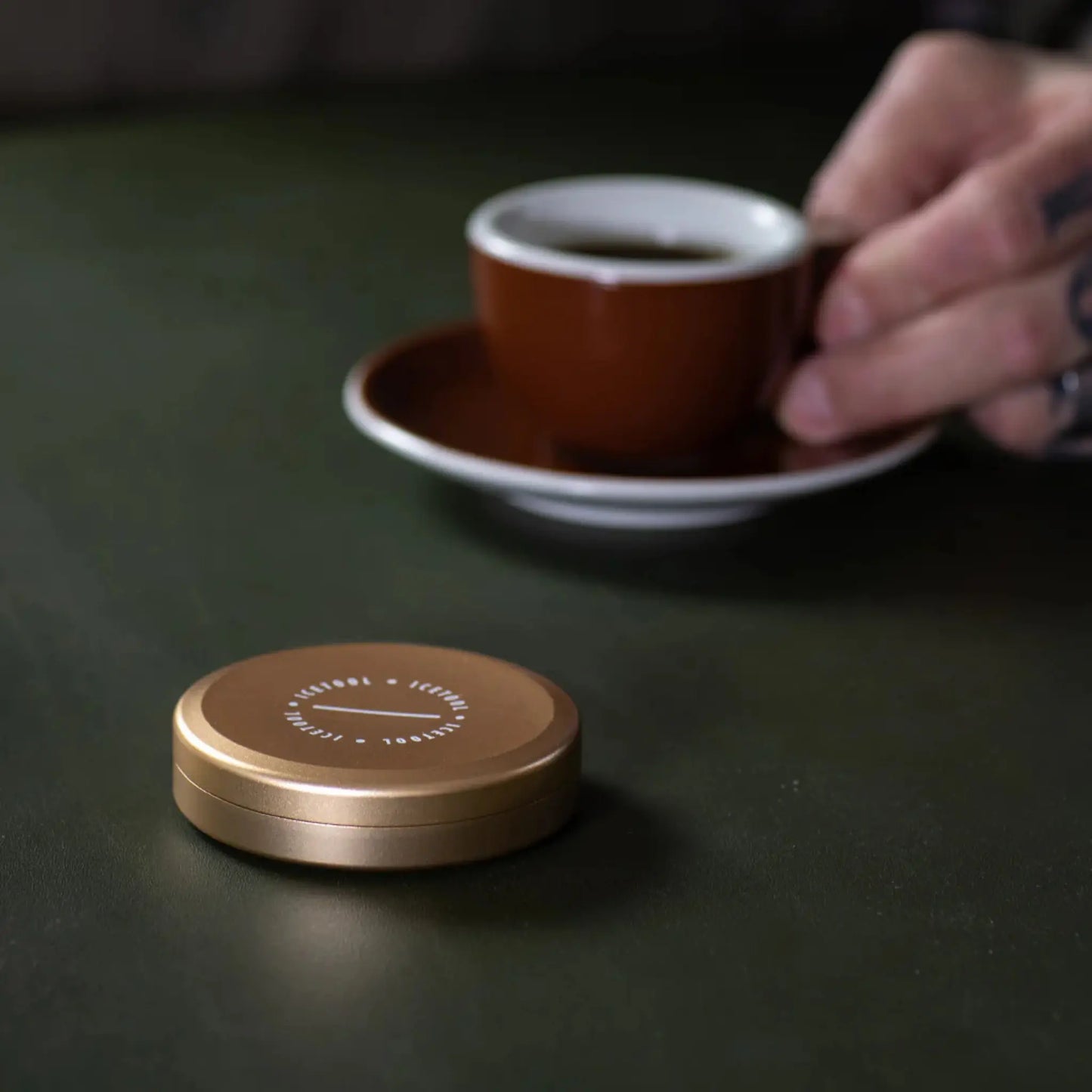 Gold coloured round metal container with Icetool branding on a dark background with a cup of coffee.